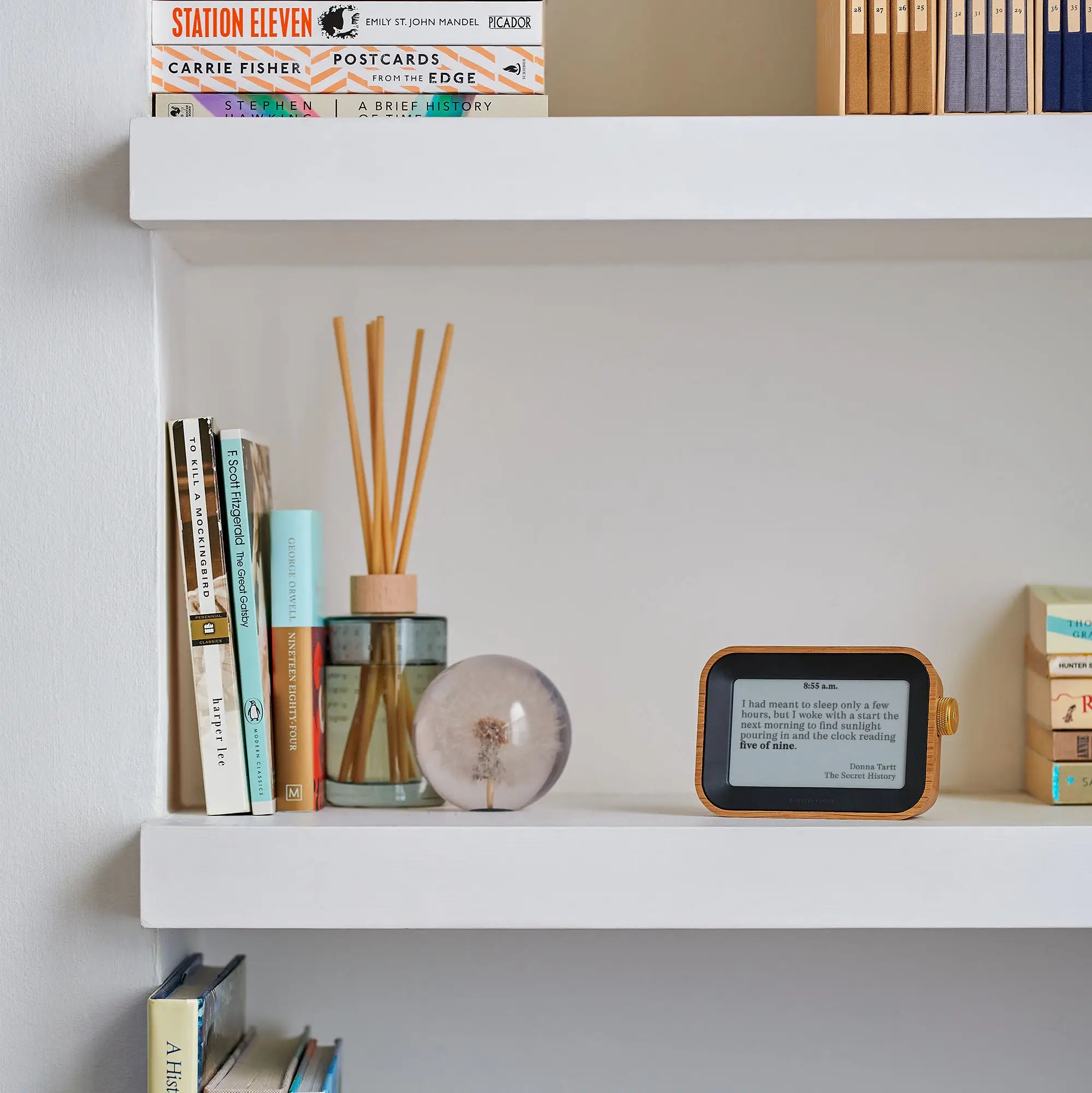 Author Clock Vol 1 Small on a white bookshelf displaying a quote from The Secret History by Donna Tartt at 8:55 a.m., beside books and a glass diffuser
