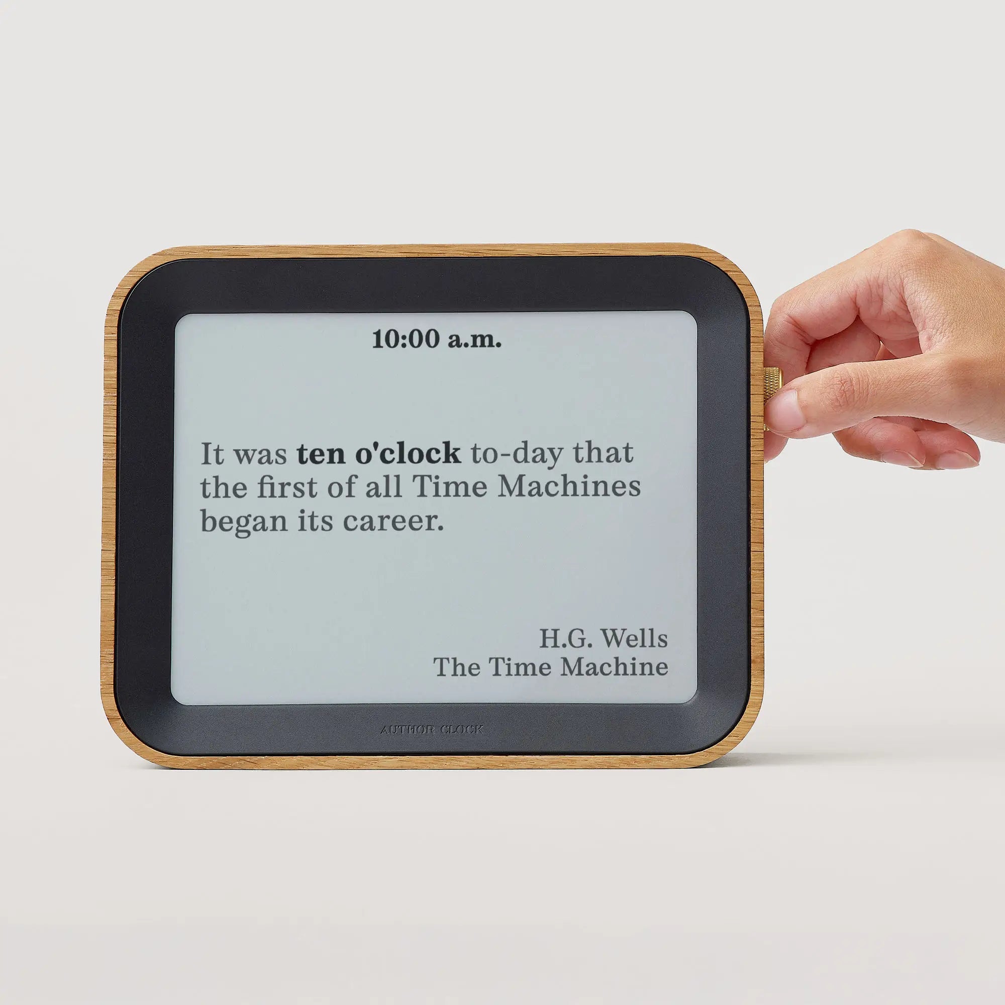 “Author Clock Vol 2 Large being adjusted by hand, showing a 10:00 a.m. display with a quote from H.G. Wells’ The Time Machine, on a white background.