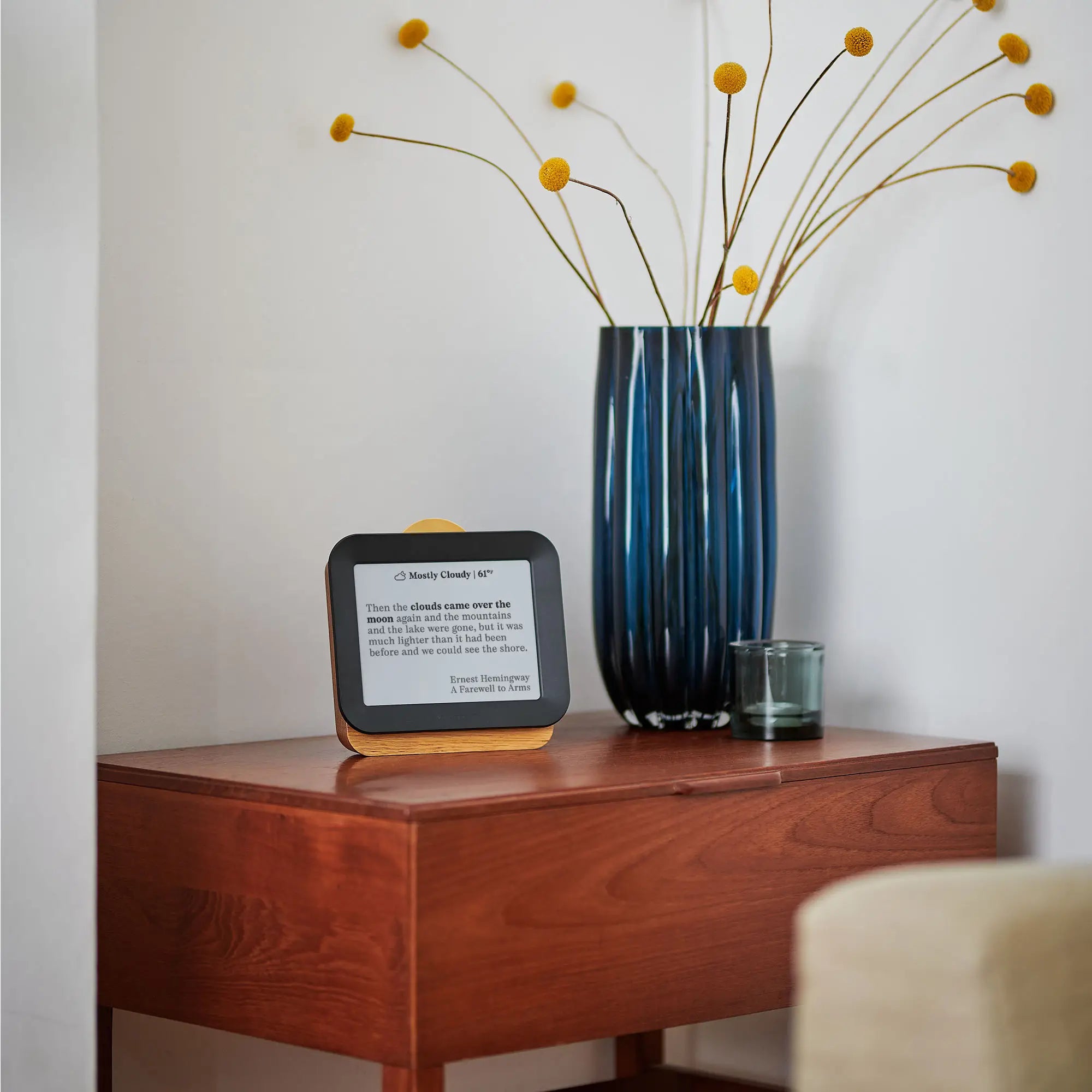 Author Forecast Vol 2 on a wooden side table displaying a quote by Ernest Hemingway from A Farewell to Arms about clouds and mountains, next to a tall blue vase with yellow flowers.